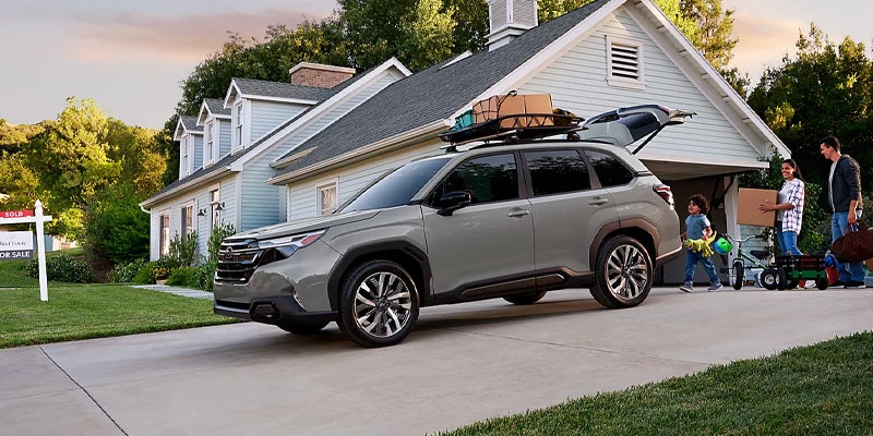 A family loads luggage into a gray SUV parked in a driveway in front of a house with a “Sold” sign