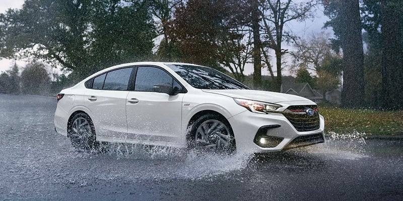 
A white Subaru Legacy sedan drives through a puddle on a rainy suburban street, splashing water