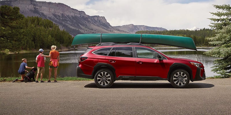 
A red Subaru Outback with a canoe on the roof is parked near a lake, with a family looking out at the water